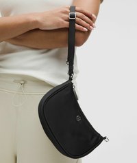 Bolso bandolera negro hecho de tela suave, con un diseño curvo, cierre de cremallera y detalles en metal plateado. La correa es ajustable.