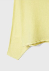 Manche et ourlet d'un pull tricoté jaune clair sur un fond gris clair uni, montrant une texture fine et un tissu doux.