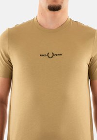 Camiseta de algodón beige con cuello redondo, con el logo "FRED PERRY" en negro centrado en el pecho, textura suave y mangas cortas.