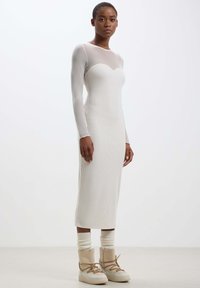 Abito a manica lunga in beige chiaro, attillato e a coste, con un corpetto in mesh trasparente e una silhouette dritta, abbinato a stivali beige con lacci.