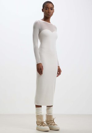 Abito a manica lunga in beige chiaro, attillato e a coste, con un corpetto in mesh trasparente e una silhouette dritta, abbinato a stivali beige con lacci.