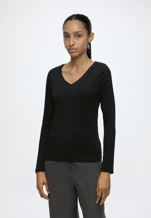 Femme aux longs cheveux foncés, portant un haut noir à col en V et manches longues et un pantalon gris, debout face à un fond clair et uni.
