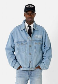 Veste en denim bleu clair avec poches avant et fermeture à boutons, ornée d'un texte brodé sur le côté gauche. Portée sur une chemise blanche avec une cravate.