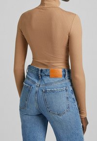 Person som bär högmidjade ljusblå raka jeans med en läderetikett och en beige ribbad polotröja, sedd bakifrån.