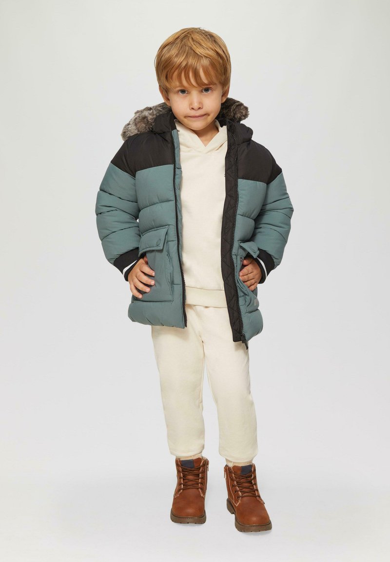 Gepolsterte Jacke in Teal und Schwarz mit fellgefütterter Kapuze, kombiniert mit einem cremefarbenen Outfit und braunen Schnürstiefeln.
