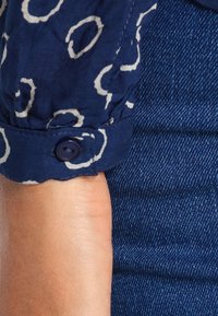 Poignet de chemise bleu marine à motifs avec des cercles abstraits blancs et un bouton foncé, associé à un tissu en denim foncé en arrière-plan.