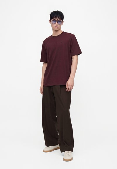 Homme portant un t-shirt violet, un pantalon large marron foncé, des baskets blanches et des lunettes de soleil teintées en bleu, debout sur fond blanc uni.