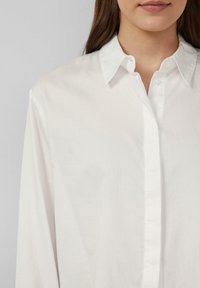Mujer con una camisa blanca de botonadura y cuello, mostrada desde los hombros hasta justo debajo de la boca sobre un fondo liso.