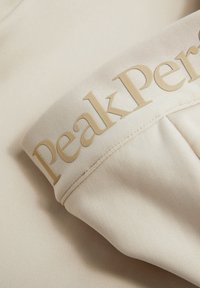 Beige sportnadrág, texturált derékpánttal, amelyen arany színű, dombornyomott 4g a "Peak Performance" felirattal. Sima anyag, minimális dizájn.