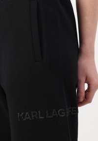 Juodos sportinės kelnės, pagamintos iš minkštos medžiagos, su šoninėmis kišenėmis ir siuvinėtu "KARL LAGERFELD" logotipu blizgia tekstūra palei koją.