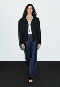 Svart texturerad blazer med en strukturerad design, ihopparad med en vit linne och mörkblå vida jeans. Svarta sandaletter med remmar och minimal smyckeskrud.