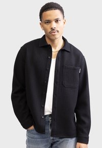 Black button-up shirt made of textured waffle fabric, featuring a chest pocket and a collar. Paired with a white undershirt and blue jeans.