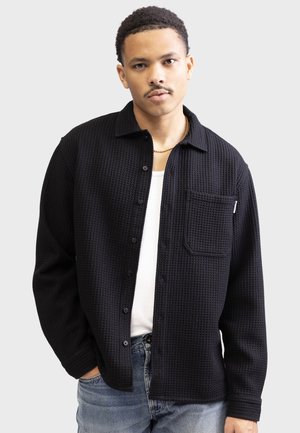 Black button-up shirt made of textured waffle fabric, featuring a chest pocket and a collar. Paired with a white undershirt and blue jeans.