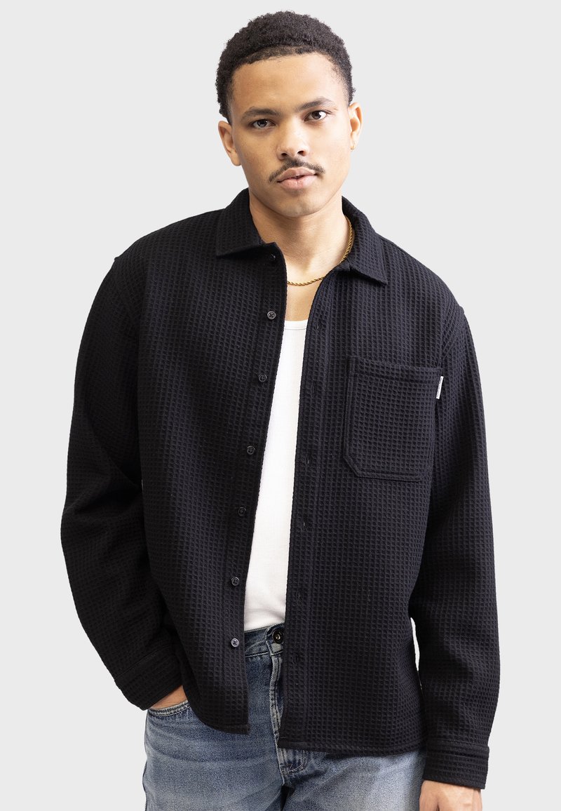 Black button-up shirt made of textured waffle fabric, featuring a chest pocket and a collar. Paired with a white undershirt and blue jeans.