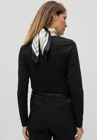 Svart långärmad topp med en slät textur, kombinerad med svarta byxor. En vit och marinblå randig silkescarf runt halsen ger detalj.