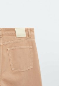 Vue arrière d'un pantalon beige montrant une poche arrière, des passants de ceinture et une étiquette rectangulaire claire sur la ceinture, sur un fond blanc.
