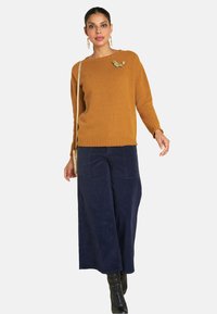 Senfgelber Strickpullover mit langen Ärmeln, kombiniert mit weiten, dunkelblauen Cordhosen, die auf der linken Seite mit einer floralen Brosche verziert sind.