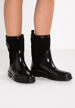 Jambes portant des bottes noires mi-mollet avec une partie inférieure en caoutchouc brillant et une partie supérieure en tissu doux, arborant un petit logo rond sur le côté extérieur.