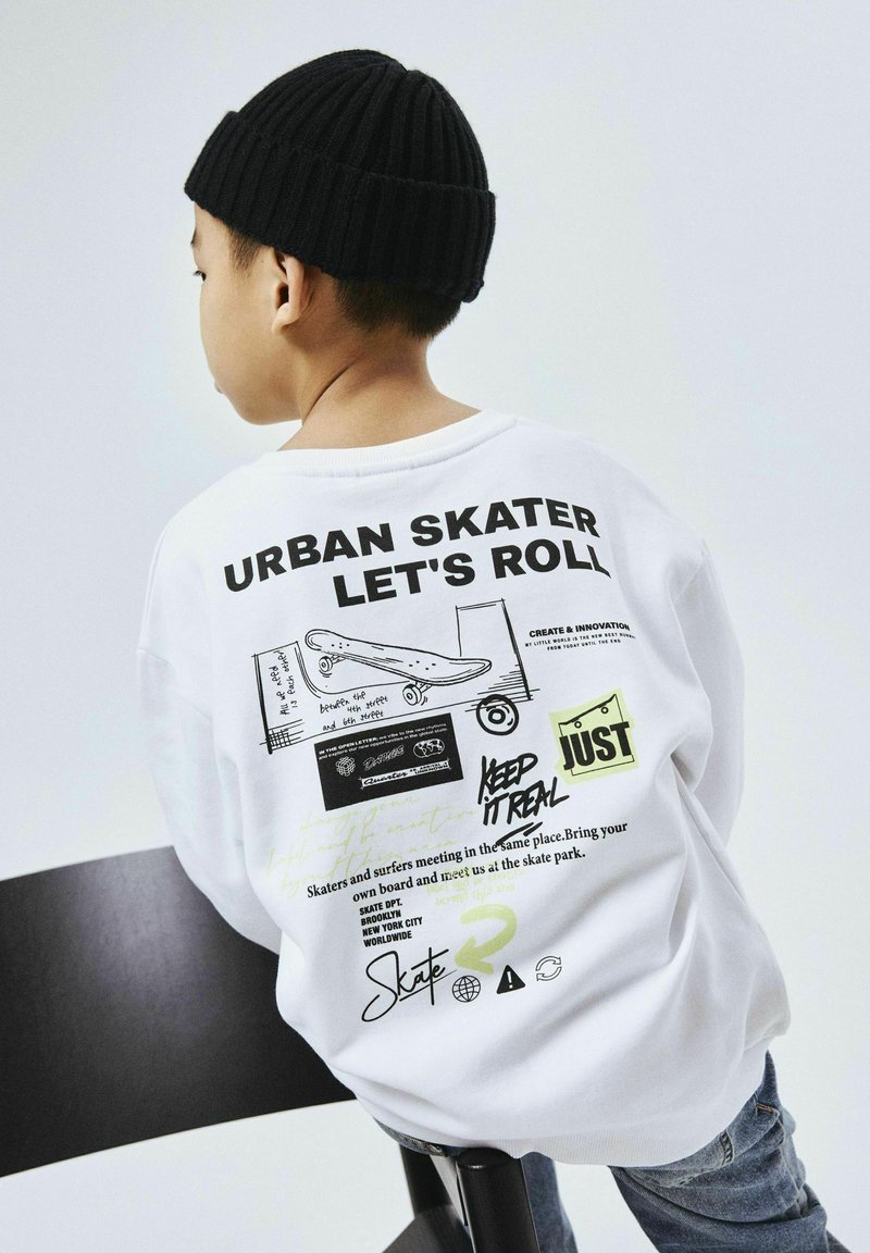 Camisa blanca de manga larga con texto y gráficos en negro, incluyendo ilustraciones de patinetas. Se lleva con un gorro de punto negro acanalado.