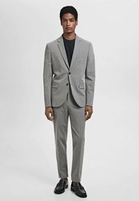 Grau Anzug mit Blazer und schmal geschnittenen Hosen, aus glattem Stoff. Schwarzes T-Shirt darunter, ergänzt durch schwarze Slipper.
