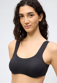 Jauna moteris su garbanotais tamsiais plaukais, dėvinti juodą berankovį liemenėlę, atsukti į priekį su neutraliu veido išraiška, paprastame šviesiame fone.