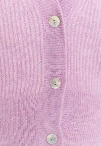 Světle růžový kardigan vyrobený z měkkého pleteného materiálu. Má žebrovanou texturu a šest kulatých, iridescentních knoflíků vpředu.
