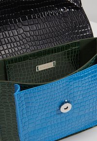 Avoin käsilaukku, jossa on kiiltävä musta, vihreä ja sininen krokotiilinkuosi sekä magneettinen suljin. Sisällä näkyy HVISK-merkintä.