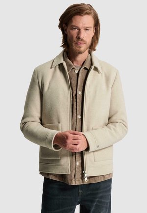 Homme aux cheveux mi-longs et à la barbe, portant une veste beige clair par-dessus une chemise marron à boutons et un jean foncé, debout, les mains jointes.