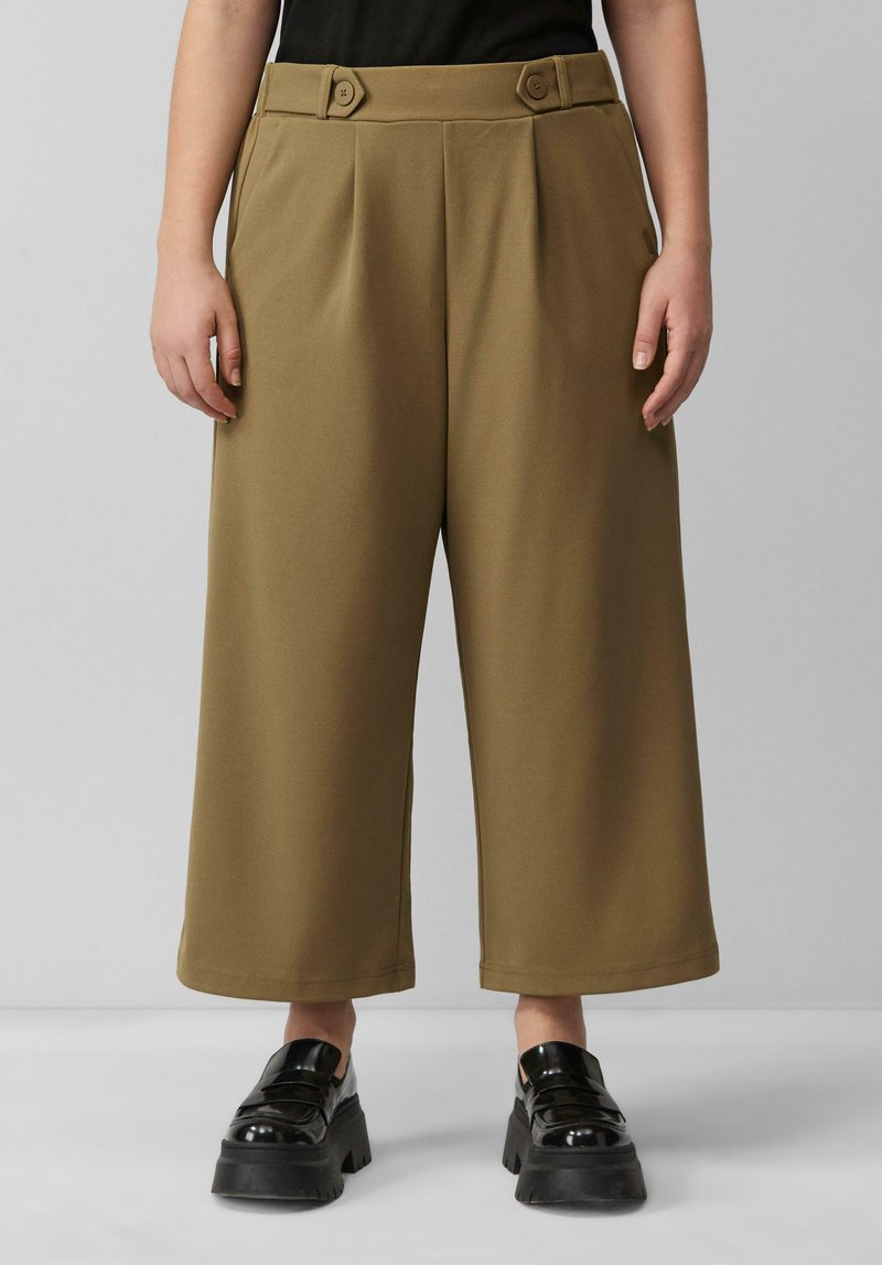 Pantalons larges marron avec des plis et des accents de boutons à la taille ; associés à des chaussures à plateforme noires et épaisses. Texture de tissu lisse.