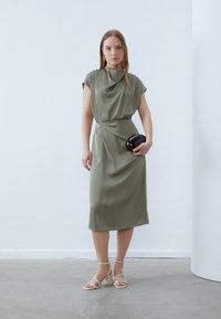 Olive green, draped midi dress with cap sleeves and a twisted waist detail. Model holds a black clutch; wearing strappy, neutral-toned heels.