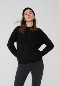 Schwarzer Kabelstrick-Pullover mit Rundhalsausschnitt und langen Ärmeln, kombiniert mit schwarzen, eng anliegenden Jeans. Die strukturierte Oberfläche sorgt für visuelles Interesse.