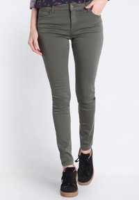 Jeans ajustados de color verde oliva con un corte elegante, cierre de botón y bolsillos frontales. Combinados con zapatillas altas negras con un acabado texturizado.
