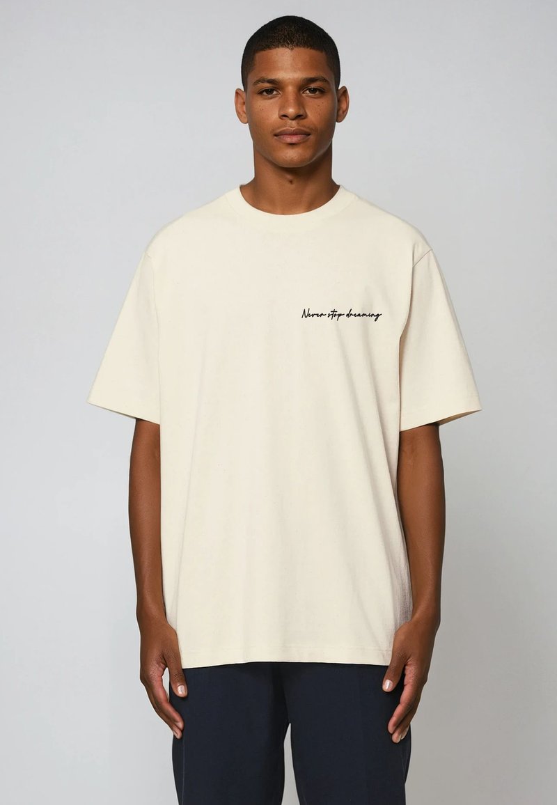 T-shirt en coton couleur crème avec une coupe décontractée. Présente des manches courtes et une impression en script noir indiquant "Never stop dreaming" sur la poitrine.