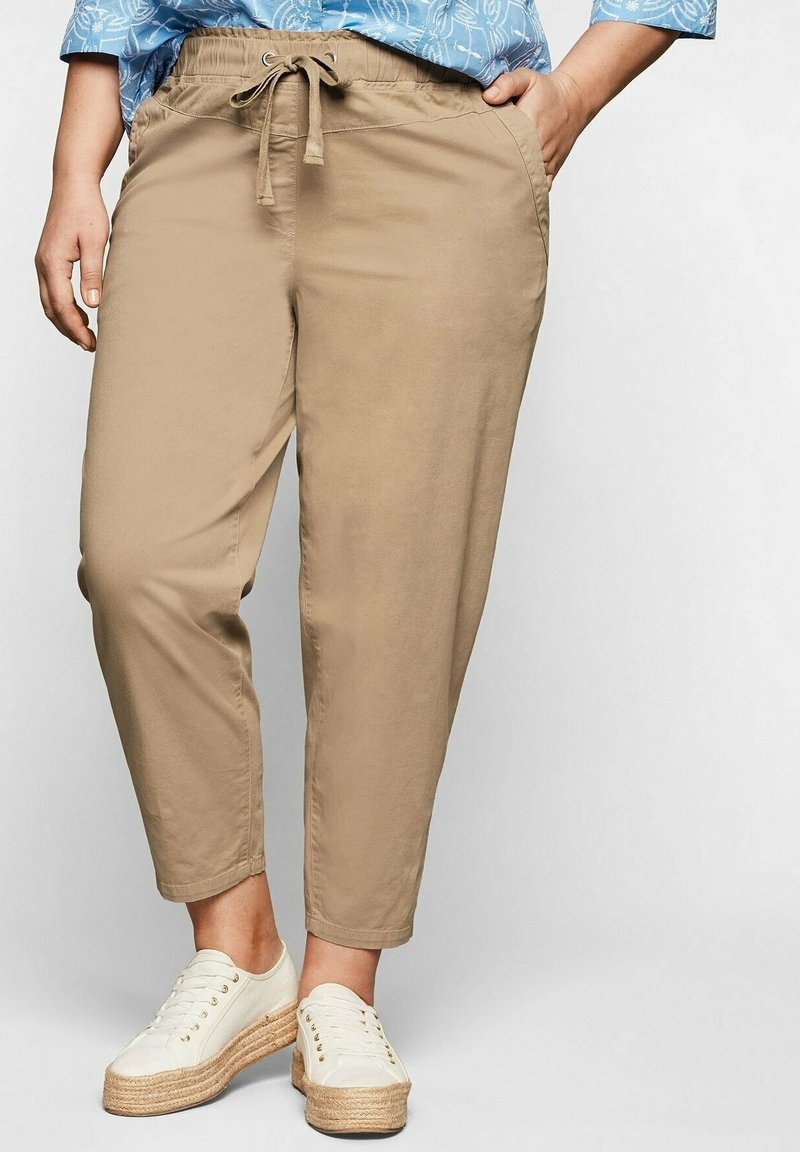 Personne portant un pantalon beige à cordon et des baskets blanches à plateforme, avec une main dans la poche, montrant une tenue décontractée du bas du corps.