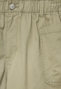 Pantalon kaki doté d'une ceinture élastique, d'une fermeture par bouton en métal, d'une poche latérale, et d'une surface en tissu lisse et texturé.