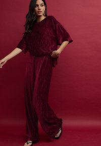 Burgundské skládání dvoudílného outfitu z měkkého sametu, s volnou blůzou a širokými kalhotami. Celkový vzhled doplňují černé sandály.