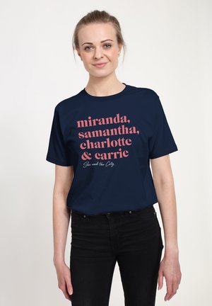 Kvinde med lys hud og samlet hår, iført en marineblå t-shirt med lyserød tekst, der viser navne fra "Sex and the City."