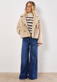 Beige dubbelzijdig jasje met zwarte stiksels, grote knopen, gedragen over een gestreept trui en wijde blauwe jeans. Glad, gestructureerd stof.