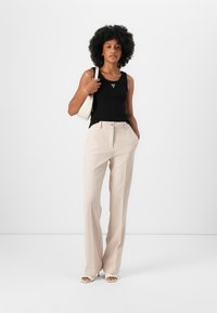 Black tank top with a triangle logo, paired with beige trousers featuring a belt and front pleats. Accessorized with a white handbag.