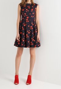 Frau in einem ärmellosen, marineblauen Kleid mit rotem Blumenmuster, plissiertem Rock und roten offenen, hässlichen Stiefeletten, die vor einer weißen Wand steht.