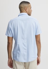 Camisa de lino de manga corta, color azul claro. Presenta un collar clásico, un corte relajado y un dobladillo curvado. Textura lisa sin patrones.