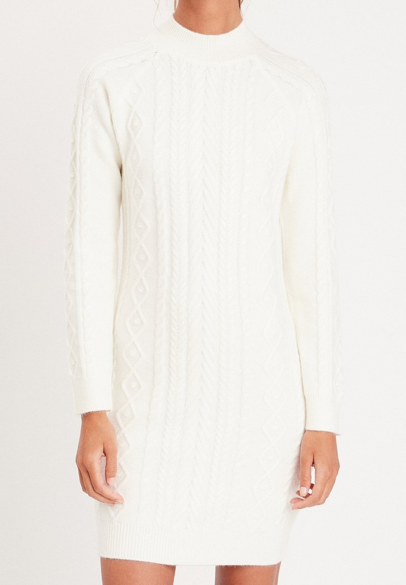 Femme portant une longue robe-pull blanche en maille torsadée, avec un col montant et des manches longues, sur un fond blanc uni.