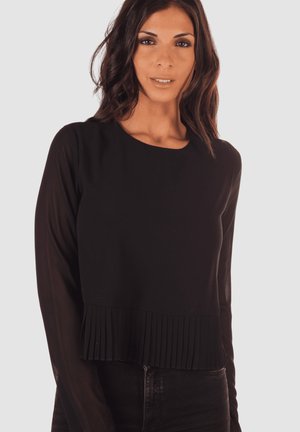 Mujer con cabello largo y oscuro que lleva un top negro de manga larga con detalles con pliegues en el dobladillo, combinado con pantalones oscuros, frente a un fondo liso.
