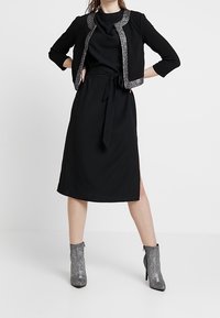 Svart klänning med en matchande kort blazer med silverdetaljer, knytning i midjan och sidoklyv, i kombination med metalliska ankelstövlar.