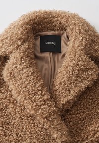 Cappotto beige texture con un tessuto esterno riccio e peloso. Presenta un ampio colletto e una fodera interna liscia. Etichetta del marchio visibile all'interno.