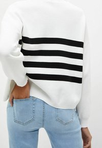 Pull blanc à rayures horizontales noires dans le dos. Texture douce, coupe décontractée, manches longues. Associé à un jean bleu clair.