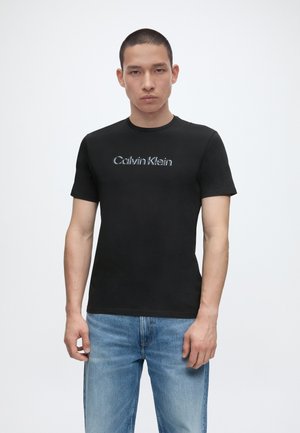 Młody mężczyzna w czarnej koszulce Calvin Klein i jasnoniebieskich dżinsach, stojący na jednolitym białym tle.