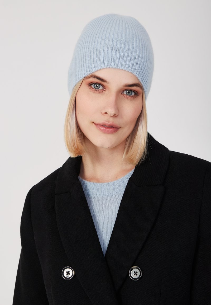 Lichtblauwe geribbelde gebreide beanie muts. Zachte textuur, comfortabele pasvorm. Gemaakt met een lichtblauwe trui en een zwarte dubbelgeknoopte jas.