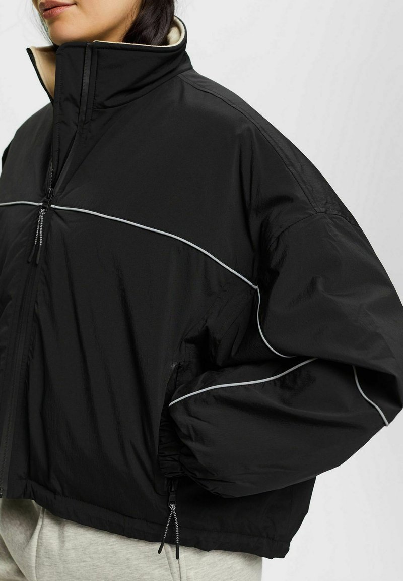 Giacca nera con colletto alto, dotata di zip frontale e dettagli in piping grigi. Realizzata in materiale leggero, con maniche ampie e voluminose.