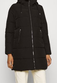 Schwarze, gesteppte Daunenjacke mit Frontreißverschluss, seitlichen Reißverschlusstaschen und Kapuze, getragen über cremefarbenen Hosen.